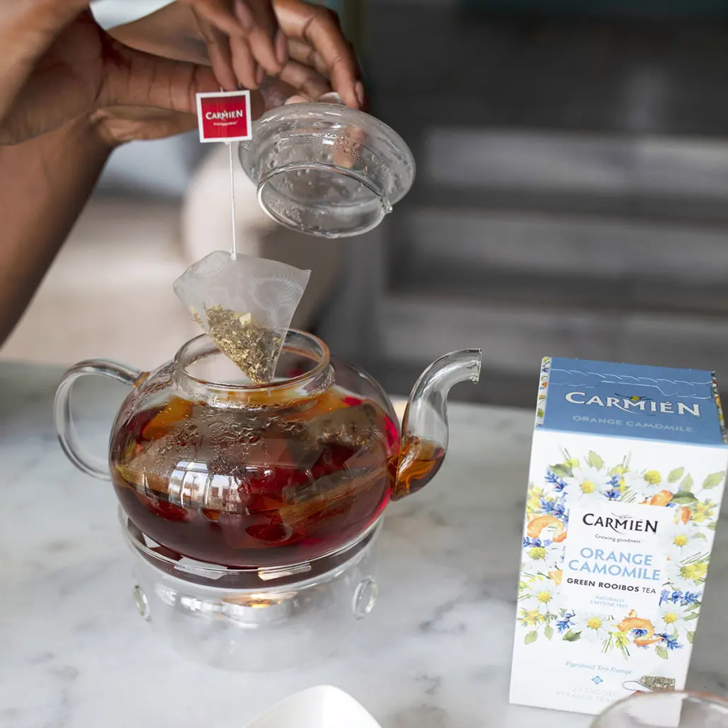 Fresh Orange Camomile Green Rooibos Tea being brewed in a clear glass teapot with a box of Carmién Tea Orange Camomile Rooibos Tea beside it, highlighting herbal tea wellness.