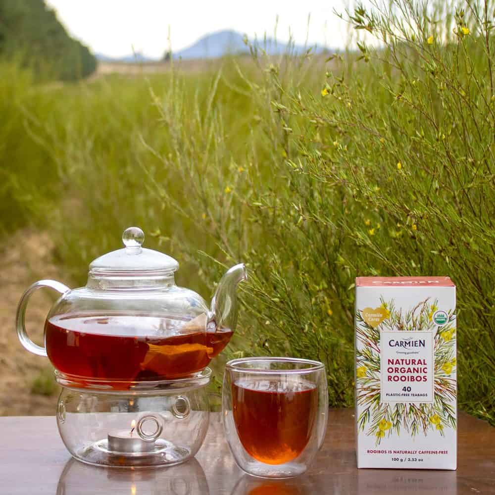 Organic Rooibos tea in a glass teapot and cup, with a box of Carmién Tea on a natural outdoor setting.