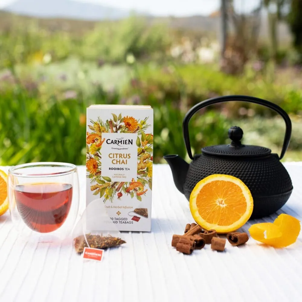 Carmién Citrus Chai Rooibos Tea with fresh orange, teapot, and tea bag on a white table outdoors.