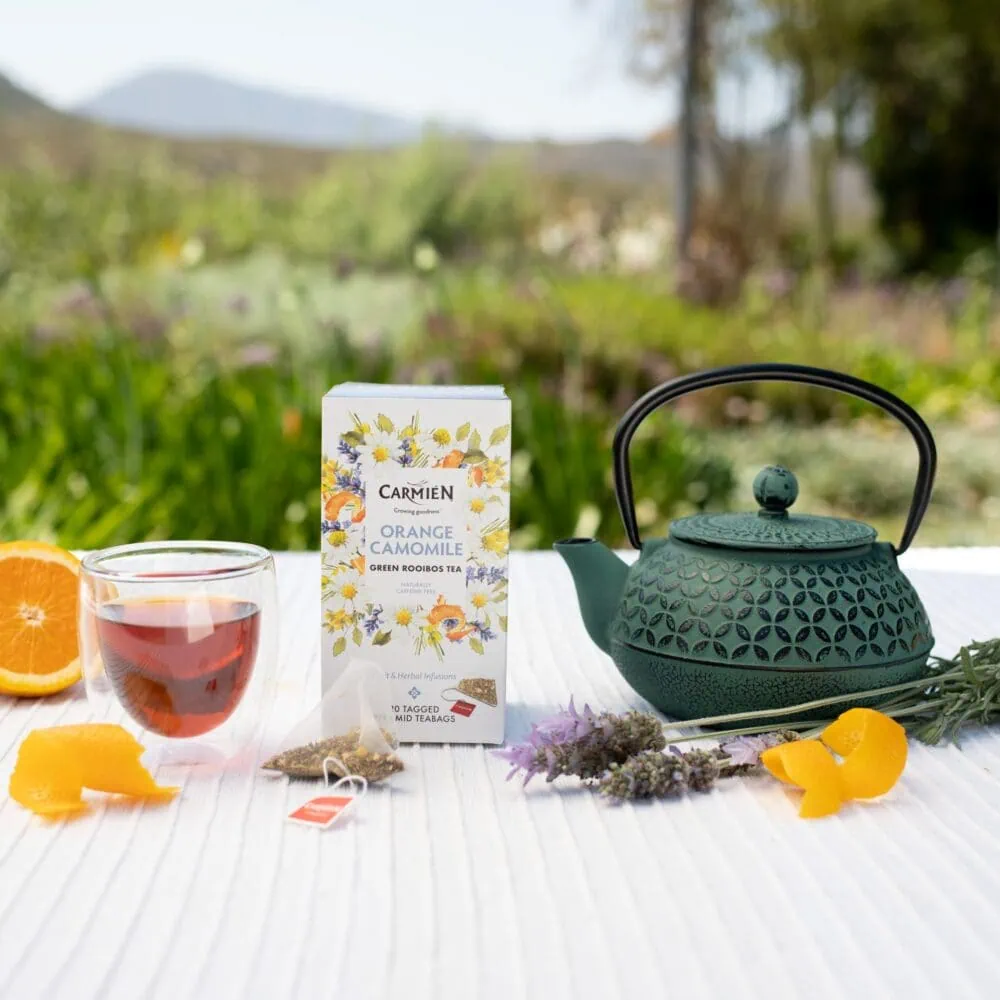 Carmién Orange Chamomile Rooibos tea in a box with a teapot and glass of tea outdoors, highlighting rooibos tea blends for health and relaxation.