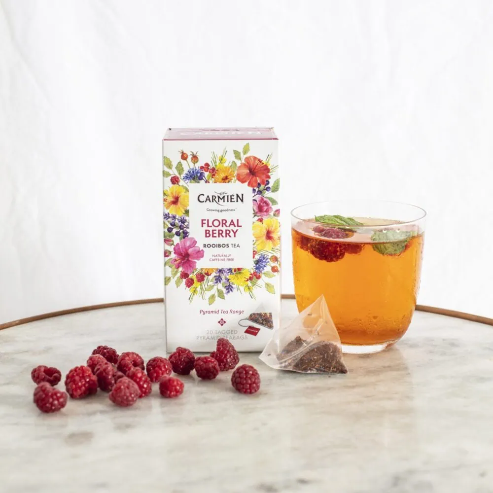 Carmién Floral Berry Rooibos tea box with a glass of brewed tea and fresh raspberries on a marble surface.