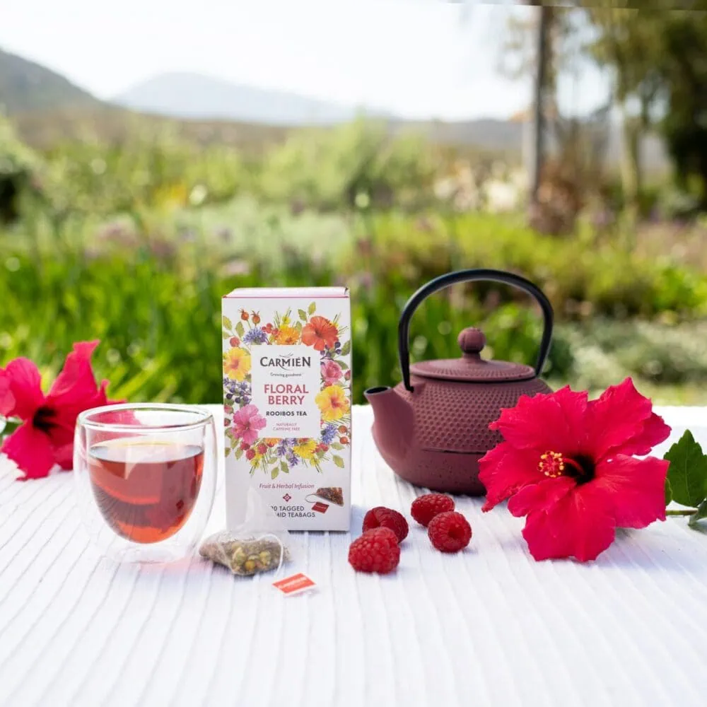 Carmién Tea Floral Berry rooibos tea with teapot, fresh raspberries, and hibiscus flowers outdoors.