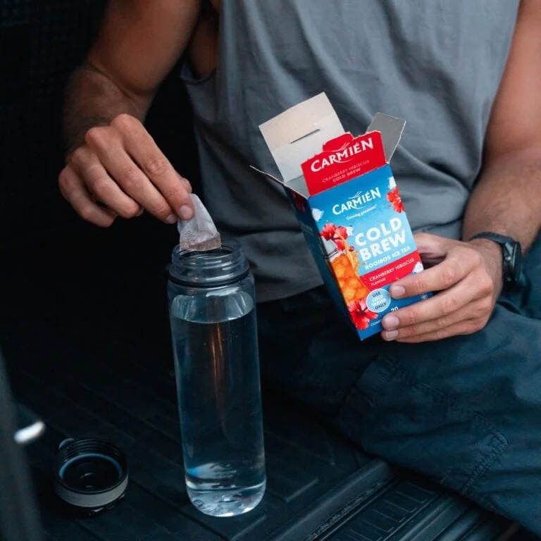 Person opening CarmiƩn Cold Brew Rooibos Ice Tea box with a teabag in a water bottle, promoting healthy rooibos tea consumption.