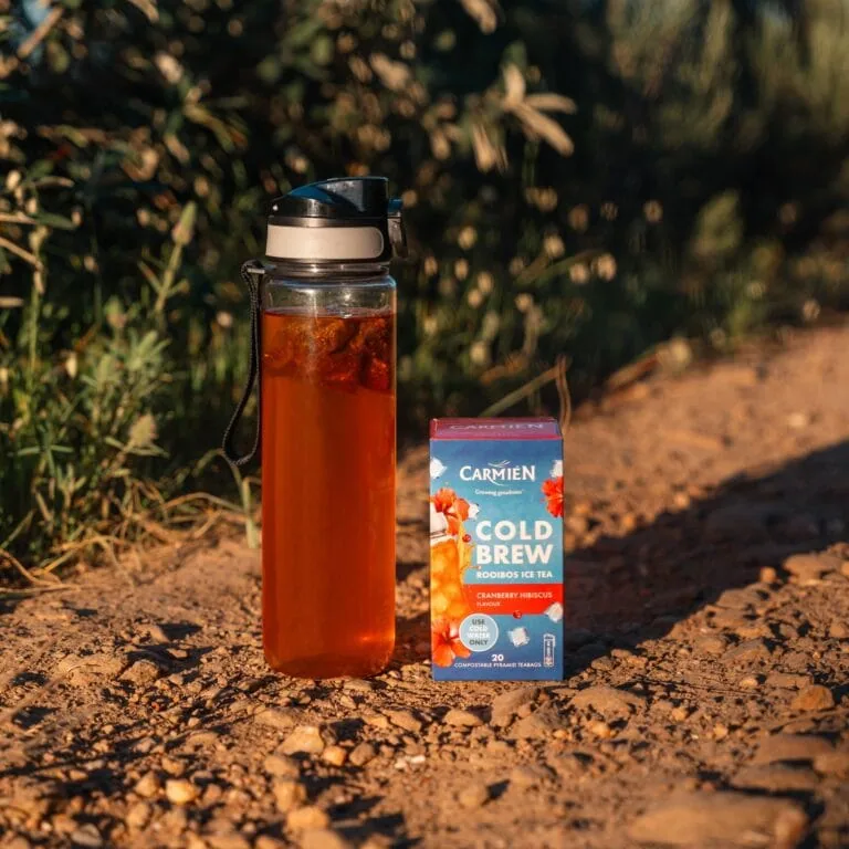 CarmiƩn Rooibos Cold Brew Tea in a glass bottle with a box of Cranberry Hibiscus flavor on a dirt path.
