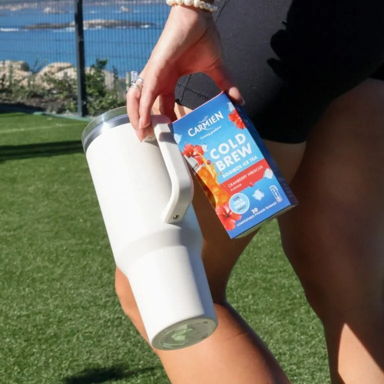 CarmiƩn Cold Brew Rooibos Ice Tea box held outdoors with a white tumbler on green grass, showcasing a refreshing tea product for e-commerce.