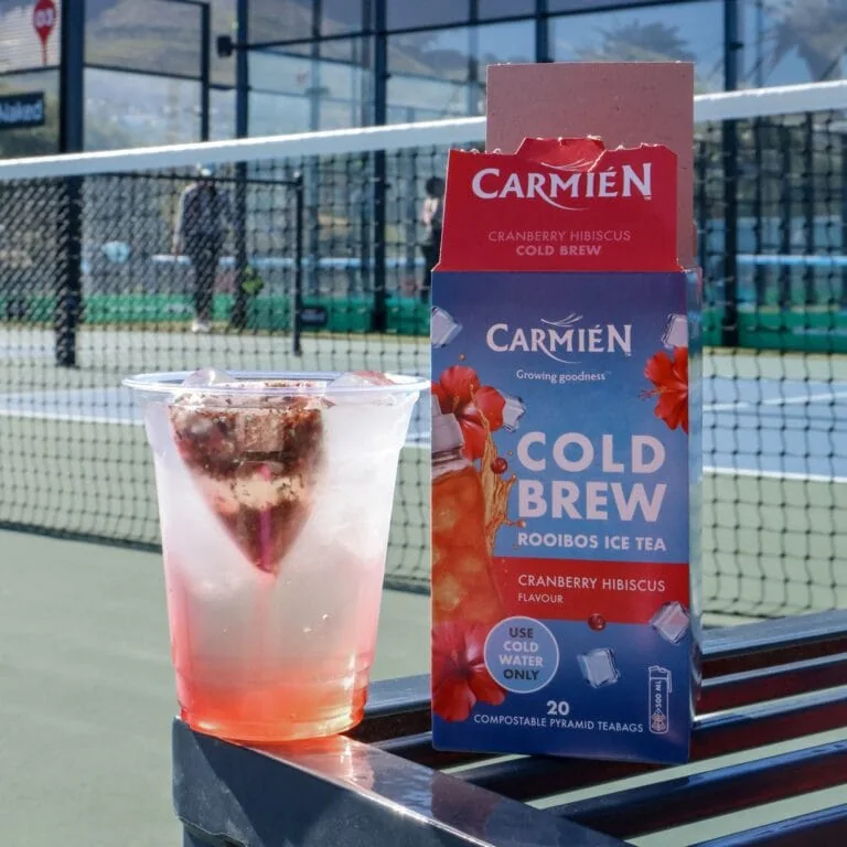 CarmiƩn Cranberry Hibiscus Cold Brew Rooibos Ice Tea box with a glass of iced tea on a tennis court.