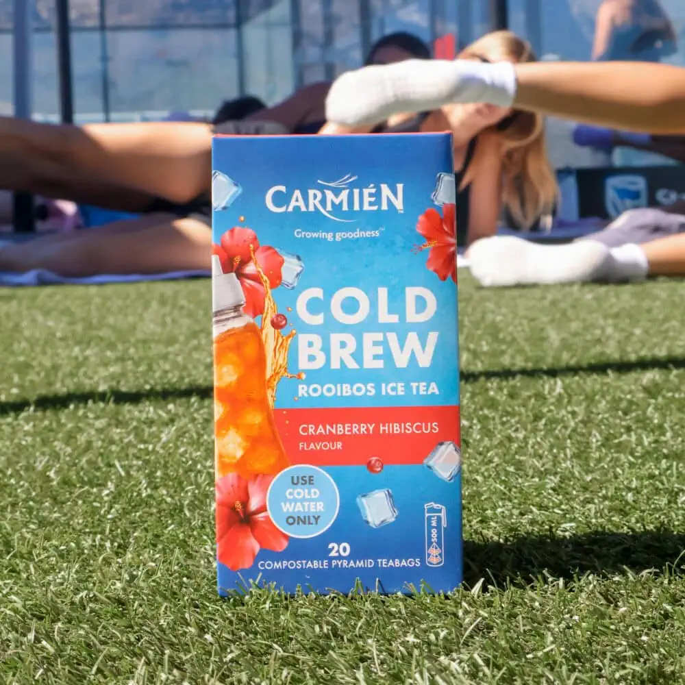 Carmién Cranberry Hibiscus Cold Brew Rooibos Tea box on grass with people stretching in the background, highlighting the refreshing tea blend for e-commerce sales.