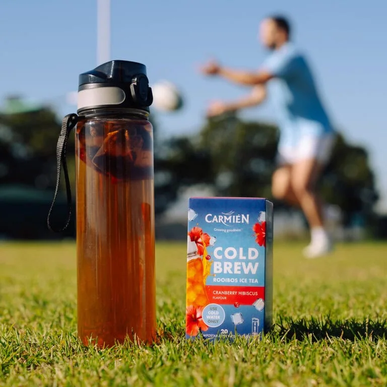CarmiƩn Cranberry Hibiscus Cold Brew Rooibos Tea in a box with a water bottle in the background, ideal for refreshing e-commerce sales.