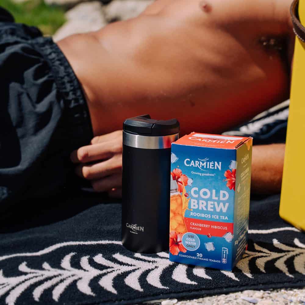 An image of a Carmién Cranberry Hibiscus Cold Brew Rooibos Tea box next to a black travel mug, set outdoors on a sunny day.