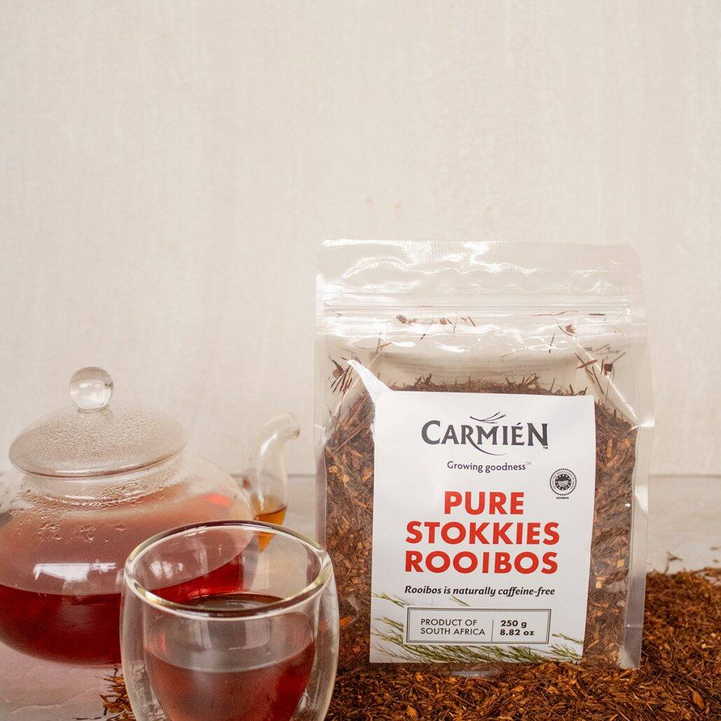 Rooibos tea in a glass cup with a sealed teapot and rooibos leaves, showcasing Carmién Tea's Pure Stokkies Rooibos, a naturally caffeine-free herbal tea from South Africa.