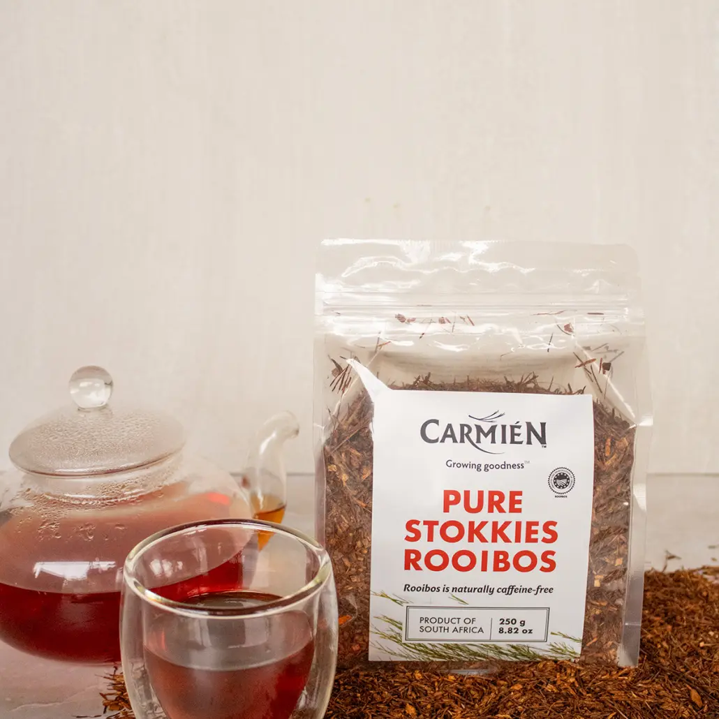 Rooibos tea in a glass cup with a sealed teapot and rooibos leaves, showcasing Carmién Tea's Pure Stokkies Rooibos, a naturally caffeine-free herbal tea from South Africa.