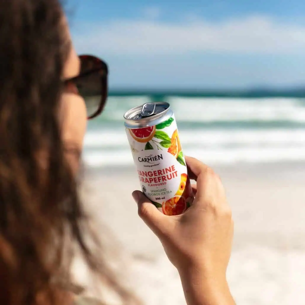 Refreshing canned Carmién Tangerine & Grapefruit Rooibos Ice Tea on the beach, showcasing a healthy, natural South African tea choice perfect for summer relaxation.
