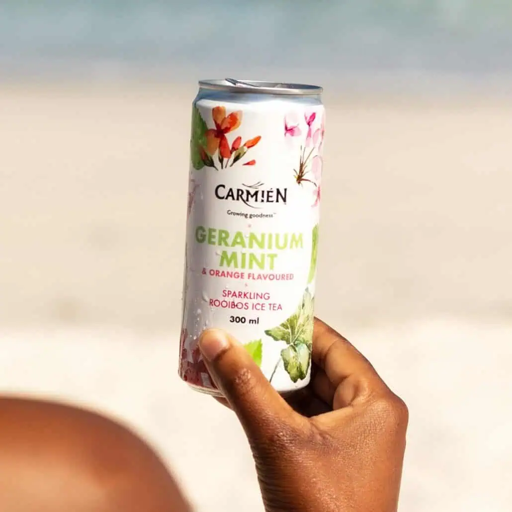 Refreshing Carmién Geranium Mint & Orange Flavoured Sparkling Rooibos Ice Tea in a 300ml can, held outdoors on a sunny beach. Perfect for cooling off during hot summer days in South Africa.