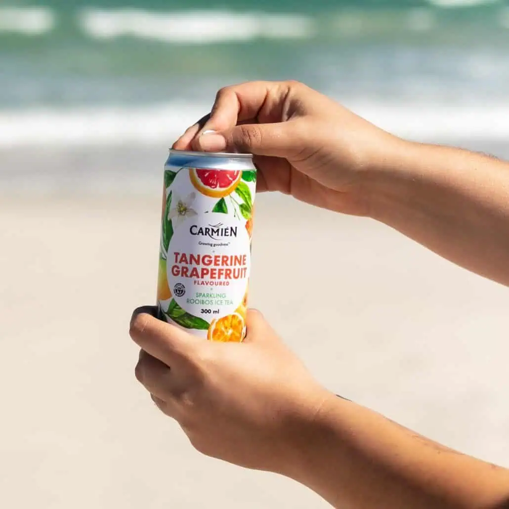 Carmién Tangerine Grapefruit Sparkling Rooibos Ice Tea in a can, held by hands on a beach background.