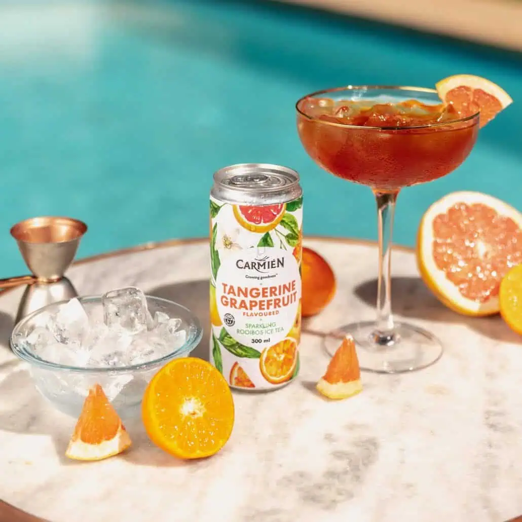 Refreshing tangerine grapefruit iced tea in a cocktail glass with citrus slices and a can of Carmién Tangerine Grapefruit flavoured Rooibos iced tea, served with ice and poolside setting.
