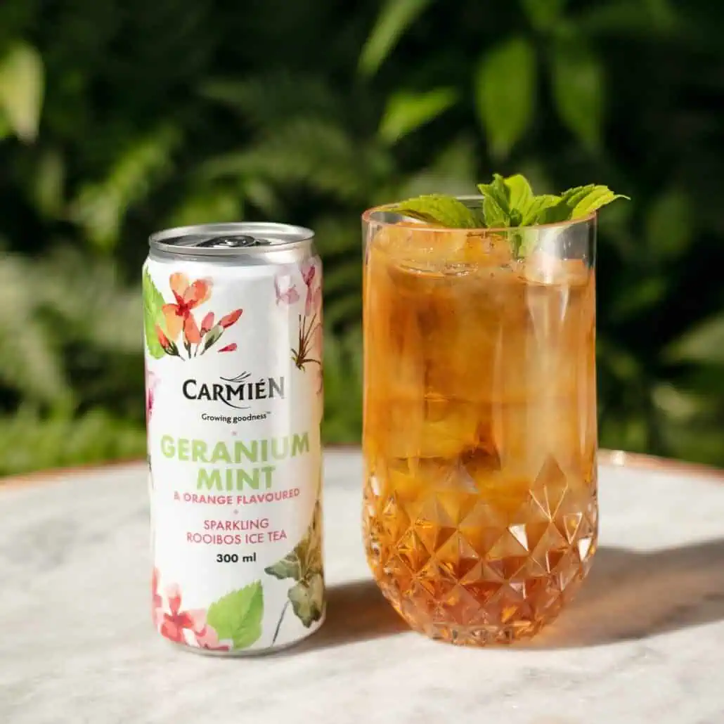 Vibrant glass of Carmién geranium mint rooibos iced tea garnished with fresh mint leaves, next to a can of flavored rooibos tea on a sunny outdoor surface.
