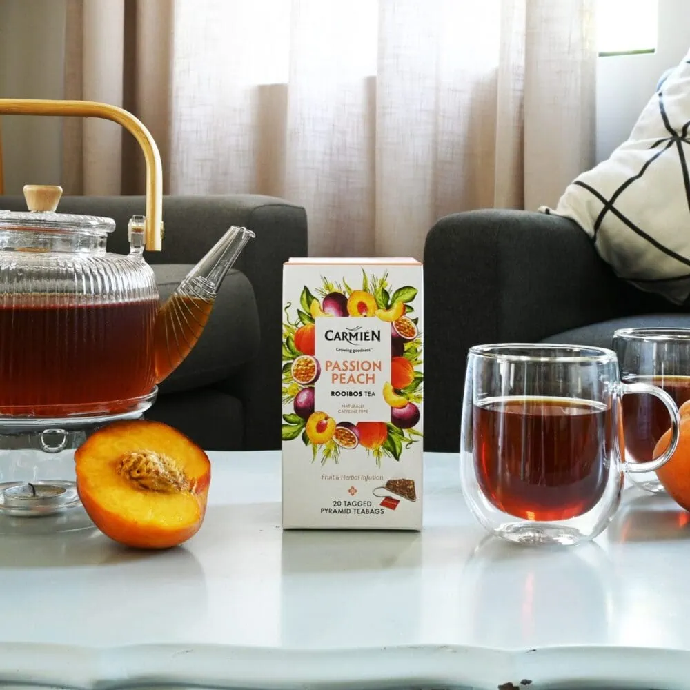 Carmién Passion Peach Rooibos Tea in a glass mug with a box of tea and a peach on a white table.