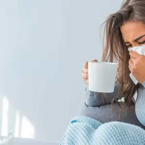 woman sitting on a couch with a cup of tea, blowing her nose with a tissue
