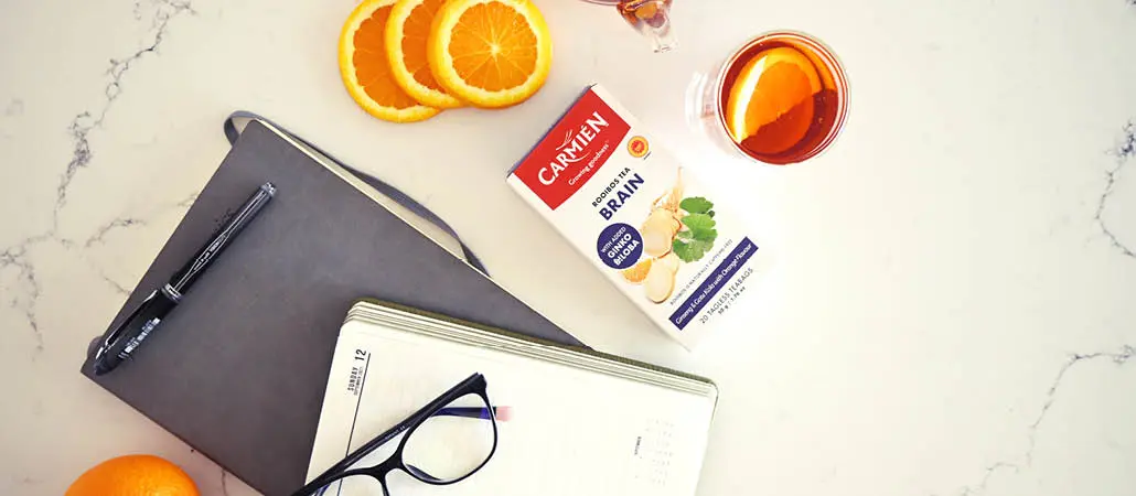 Carmién Tea with herbal ginger immune booster, orange slices, and a glass of brewed tea on a white marble surface, highlighting natural ingredients and wellness benefits.