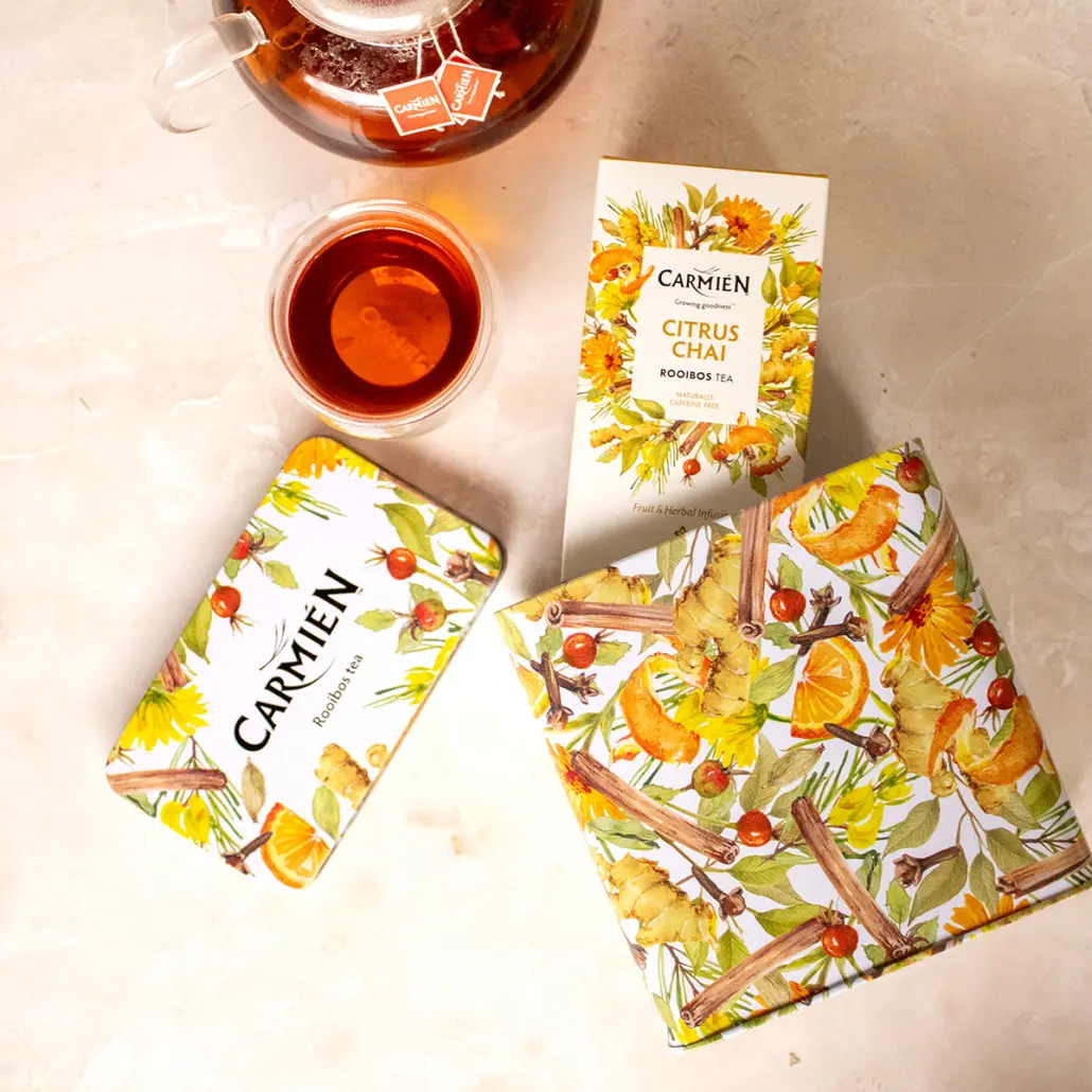 Carmién Citrus Chai Rooibos Tea in vibrant packaging with fresh herbal ingredients, steeps in a glass teapot and cup, showcasing a natural, herbal, and citrus-infused tea experience.