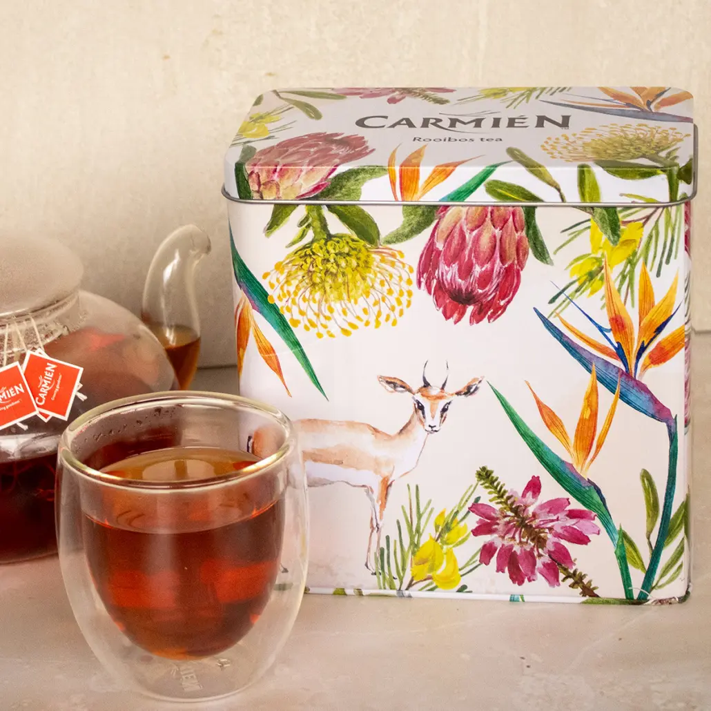 Rich rooibos tea in a double-walled glass cup with a colorful Carmién Tea tin featuring floral and wildlife illustrations, showcasing premium South African herbal tea blends for wellness and relaxation.