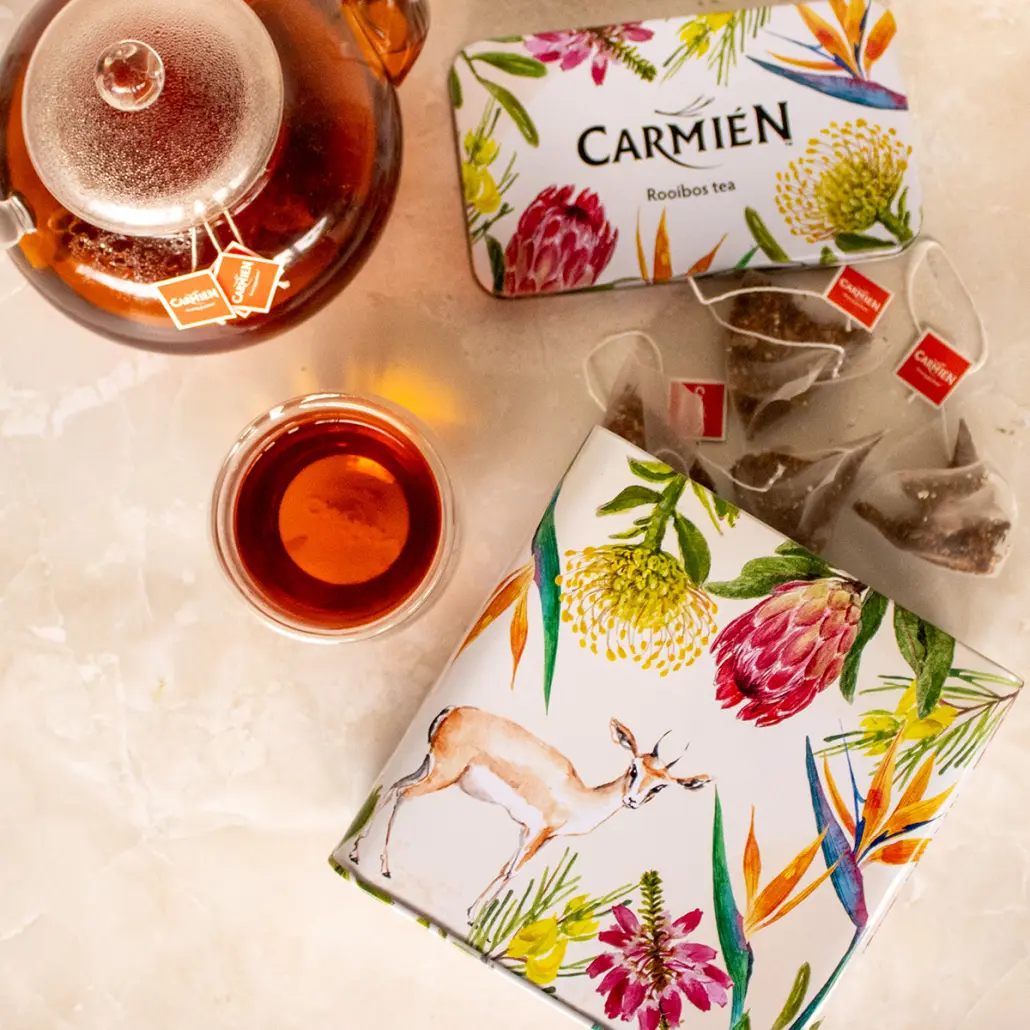 Rich Rooibos tea in a glass teapot with Carmién Tea branded tea bags, accompanied by herbal tea infusions in decorative packaging, showcasing vibrant botanical illustrations and traditional South African tea culture.