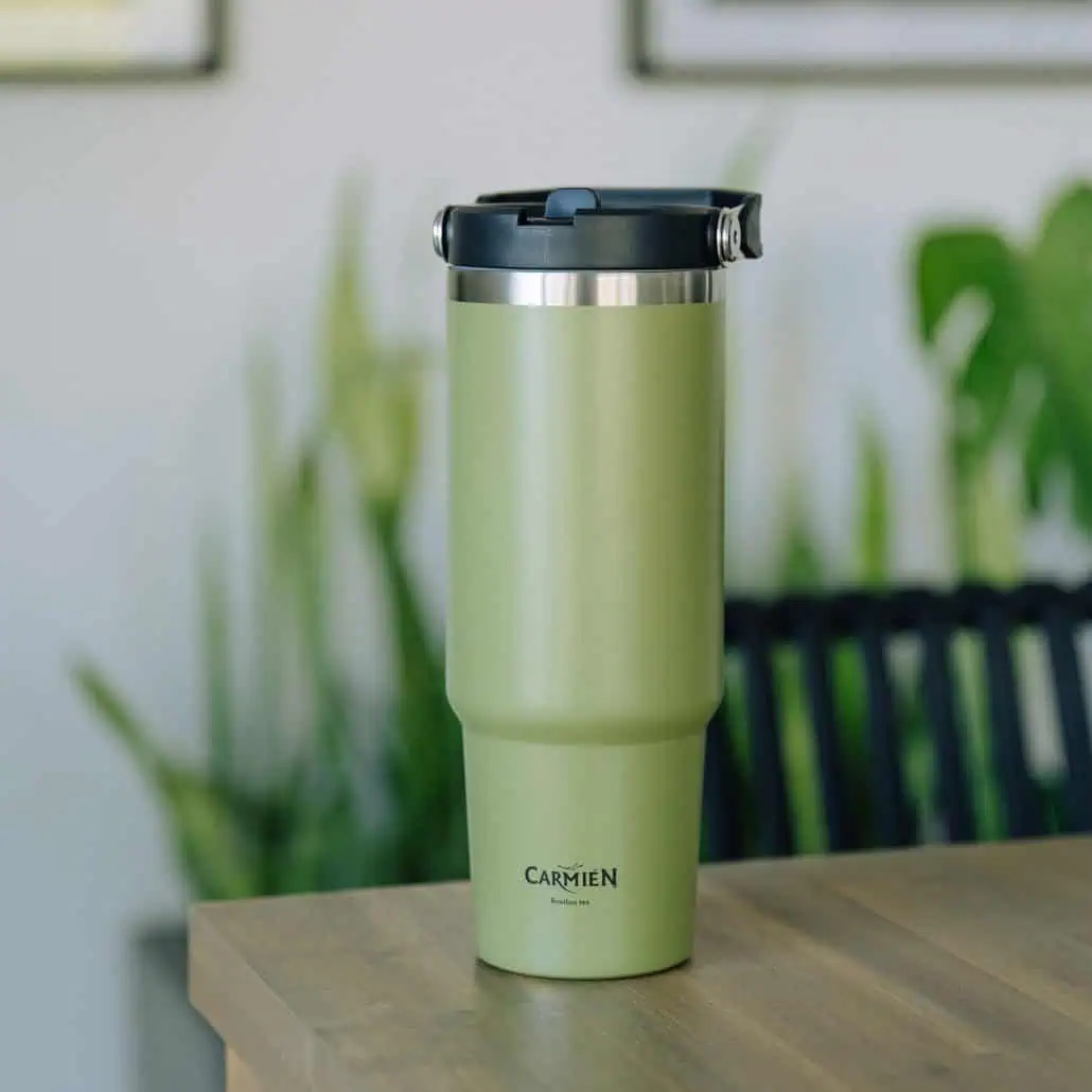 Sustainable green Carmién Tea tumbler on wooden table with lush green plants in the background, ideal for hot and cold tea, promoting healthy lifestyle.