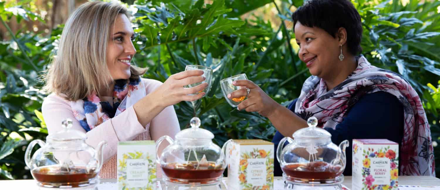Rich, aromatic Carmién Tea herbal infusions enjoyed outdoors in lush greenery with women toasting their tea glasses. Authentic South African herbal teas offer wellness and unique flavours.