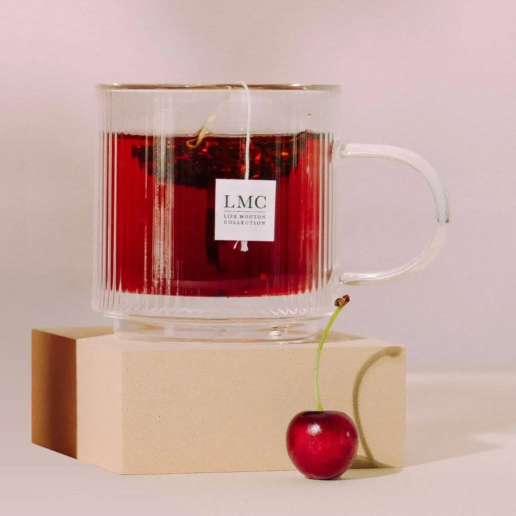 Bright red Carmién herbal tea in a clear glass mug with a cherry next to it on a beige block, showcasing a vibrant, natural beverage made from South African wild-harvested herbs.
