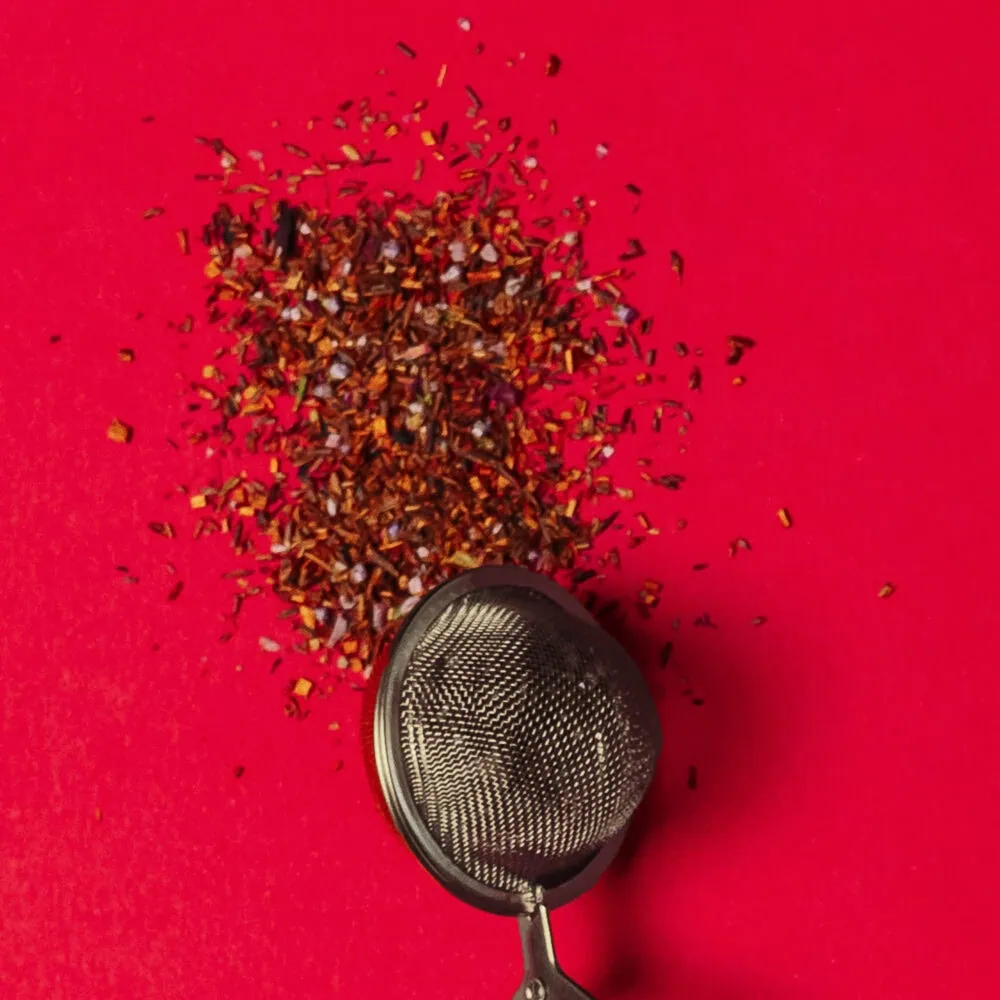 Loose Carmién Tea leaves spilled on a vibrant red background with a metal tea infuser. Perfect for herbal tea lovers seeking authentic South African rooibos tea.