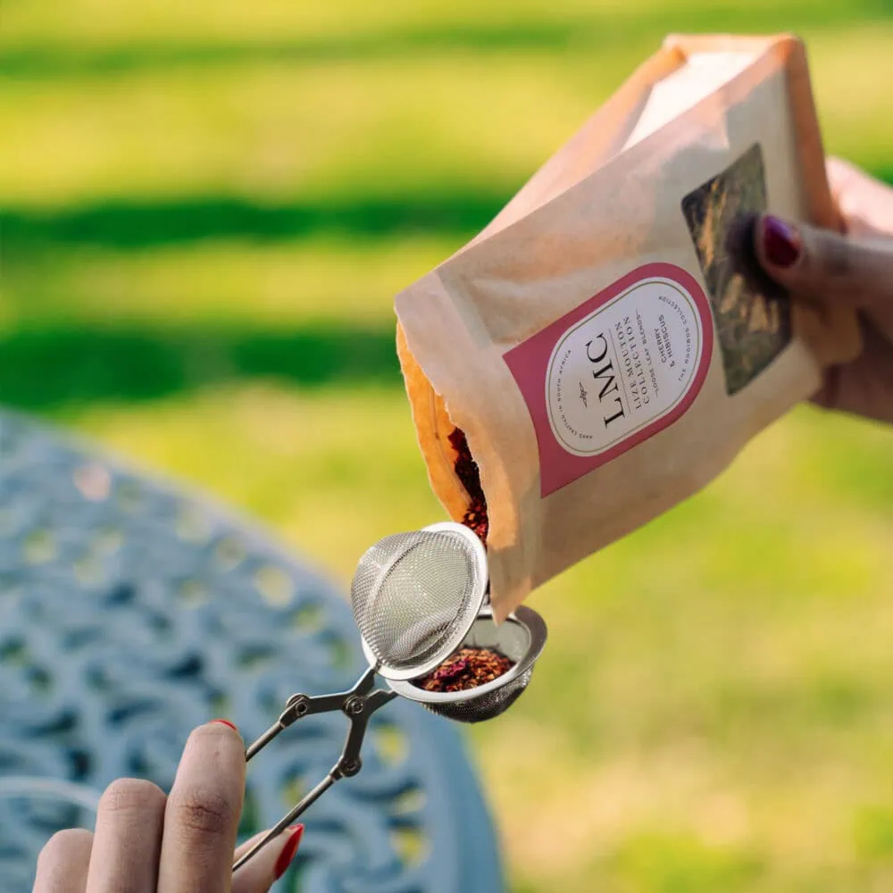 Hand pouring loose Rooibos tea from a paper bag into a tea infuser outdoors, showcasing natural herbal tea preparation.