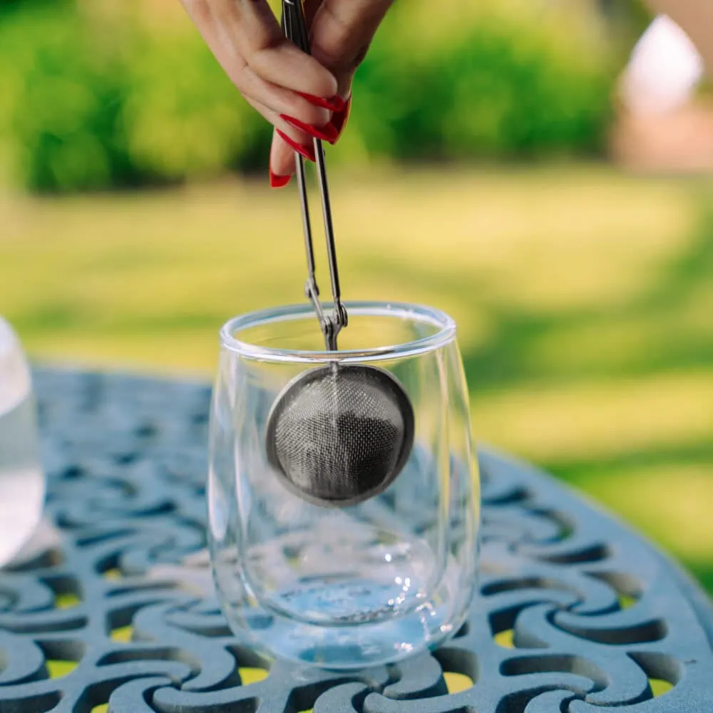 Hand using a tea infuser to brew Carmién Tea in a clear glass, outdoors setting with a blurred green background. Perfect for enjoying natural herbal tea.