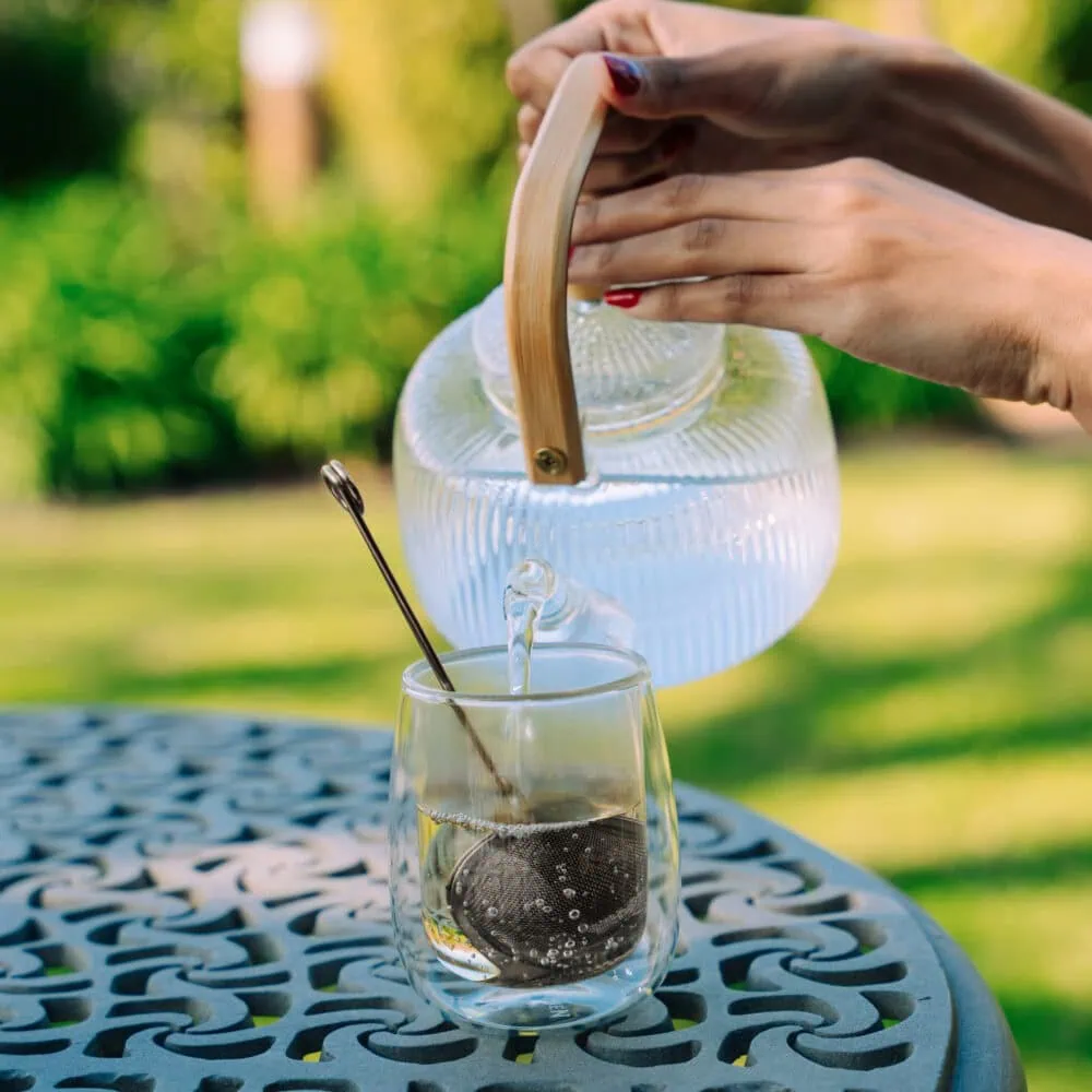 Hand pouring hot herbal tea from a glass teapot into a glass cup outdoors, showcasing natural ingredients and traditional brewing methods.