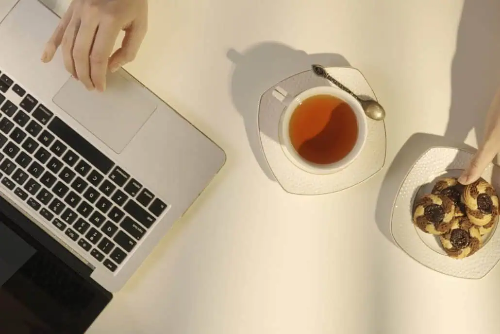 A person enjoying a cup of Carmién Rooibos Tea alongside a plate of cookies and a laptop, highlighting a healthy alternative to coffee for avoiding the 3pm crash.