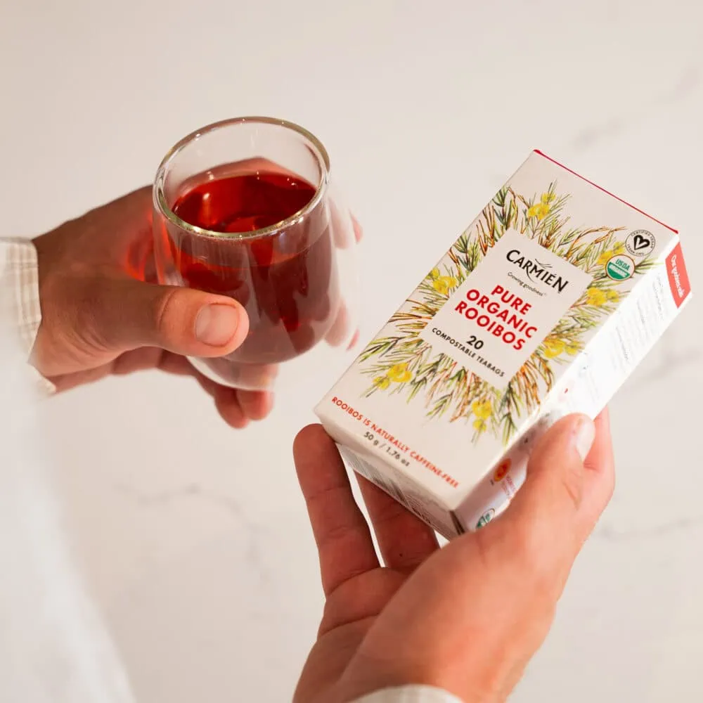Hand holding a glass of brewed organic Rooibos tea next to a box of Carmién Tea's Pure Organic Rooibos (20 pillow teabags), highlighting natural, healthy herbal tea.