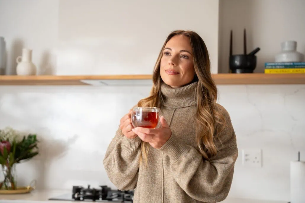 Rich, warm Carmién Tea in a glass cup, held by a woman in a cozy kitchen setting, highlighting natural herbal tea benefits and South African heritage. Perfect for health-conscious tea lovers.