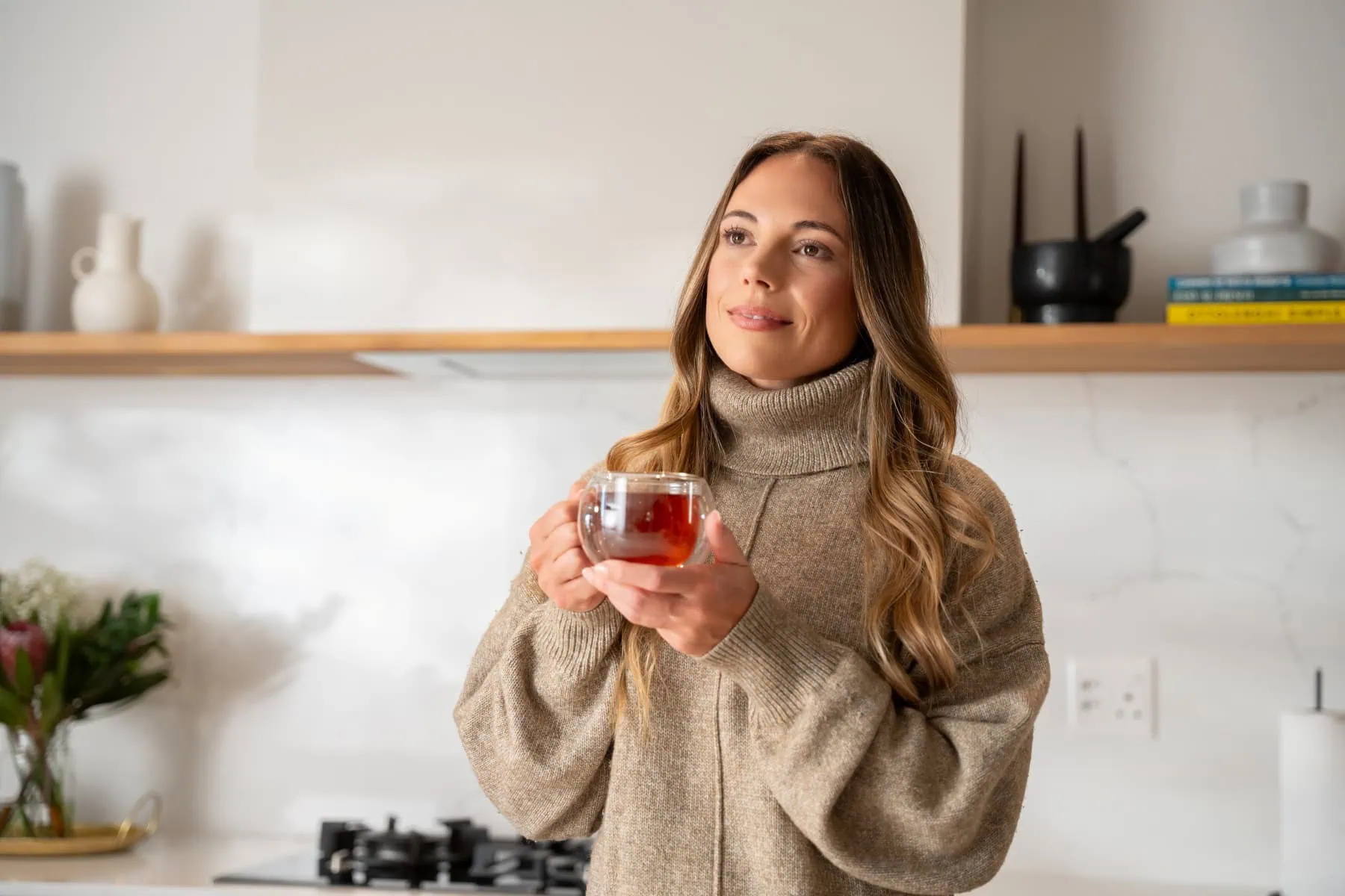 Rich, warm Carmién Tea in a glass cup, held by a woman in a cozy kitchen setting, highlighting natural herbal tea benefits and South African heritage. Perfect for health-conscious tea lovers.