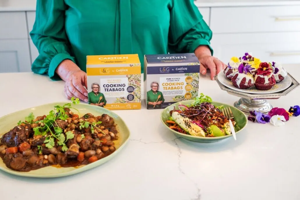 Carmién Tea boxes displayed with homemade desserts and a savory dish on a white kitchen counter, highlighting rooibos tea blends for health and wellness.