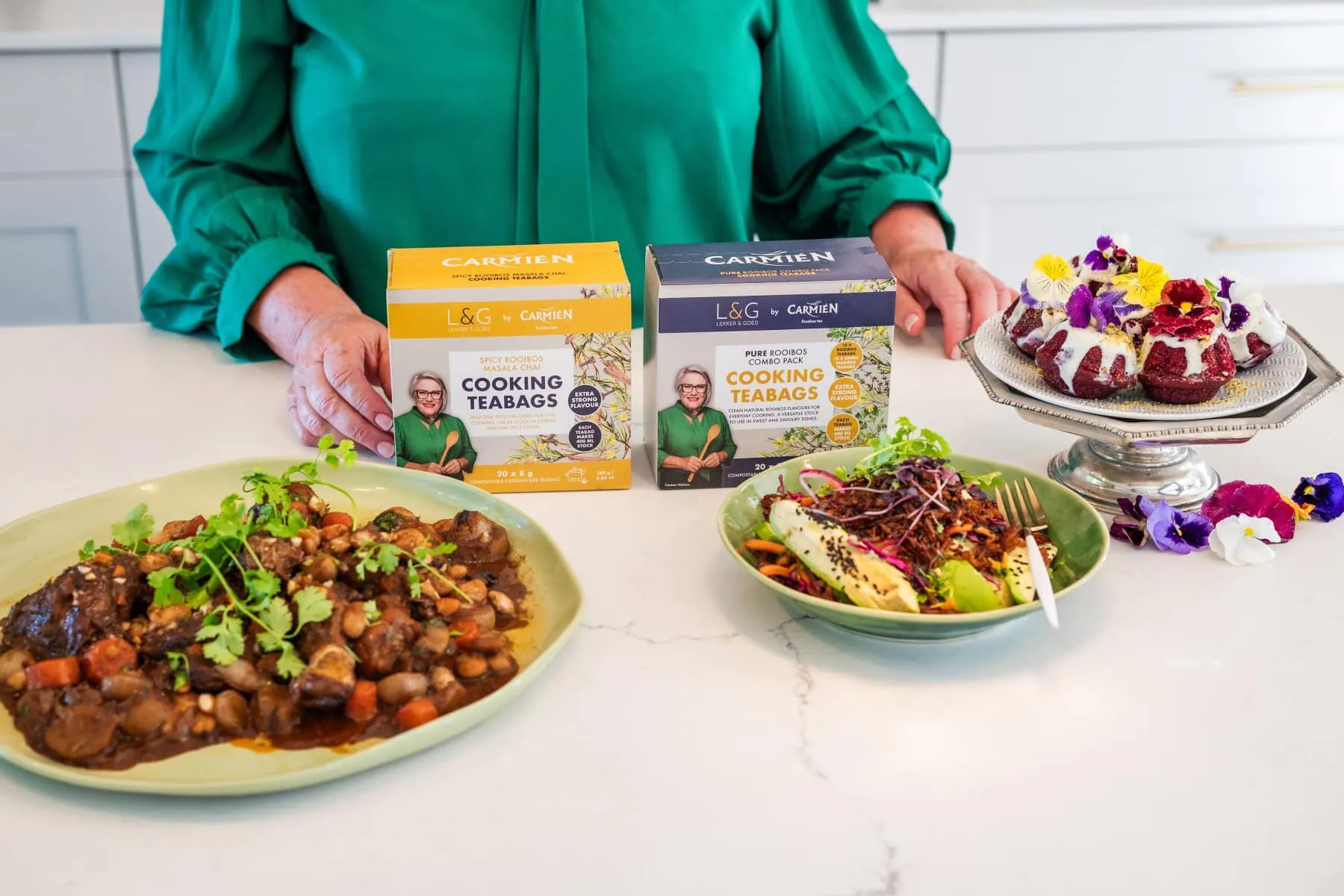 Carmién Tea boxes displayed with homemade desserts and a savory dish on a white kitchen counter, highlighting rooibos tea blends for health and wellness.