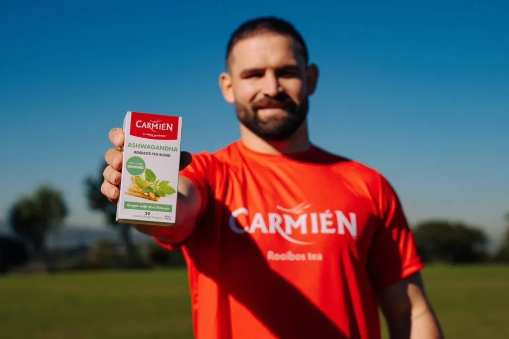 Man in red Carmién Tea shirt holding a box of Rooibos tea outdoors with a clear blue sky.
