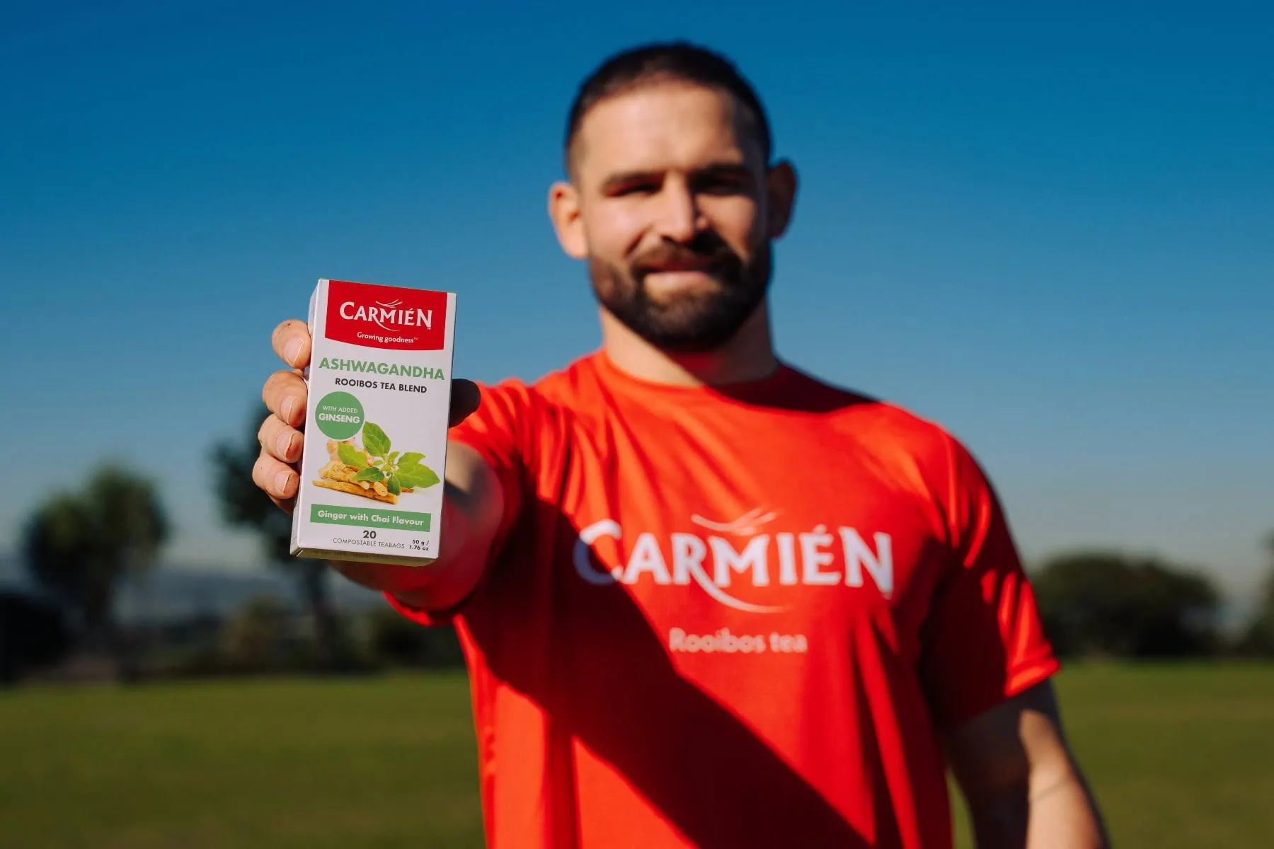 Man in red Carmién Tea shirt holding a box of Rooibos tea outdoors with a clear blue sky.