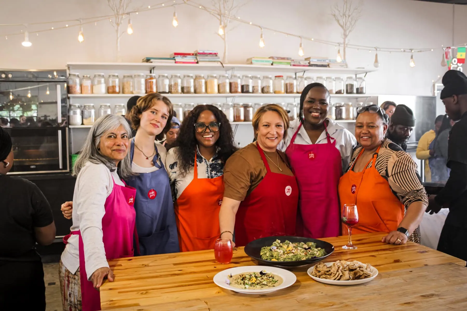 Diverse group of women in aprons smiling in a tea shop with rooibos tea blends and snacks on a wooden counter.